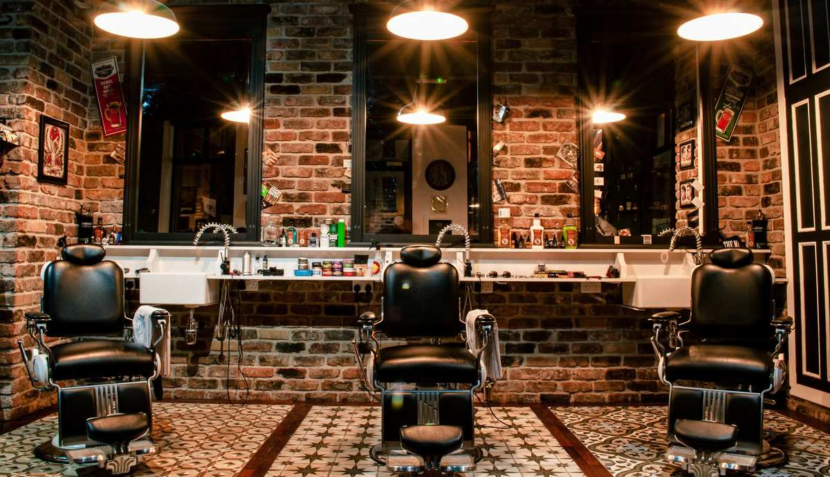 Three black leather barber chairs in front of mirrors and a brick wall, lit by warm pendant lamps.