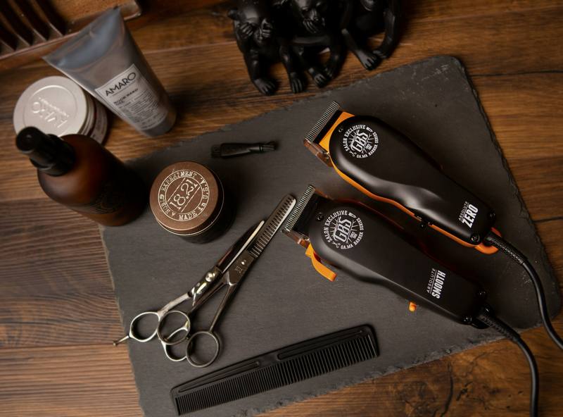 Two hair clippers, scissors, a comb, and grooming products laid out on a slate board.